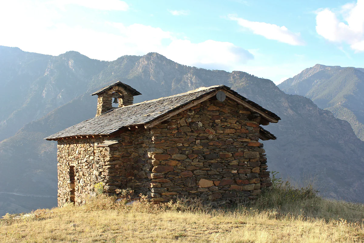 La capilla de Sant Romà d'Auvinyà, de piedra, sobre un prado seco con las montañas detrás