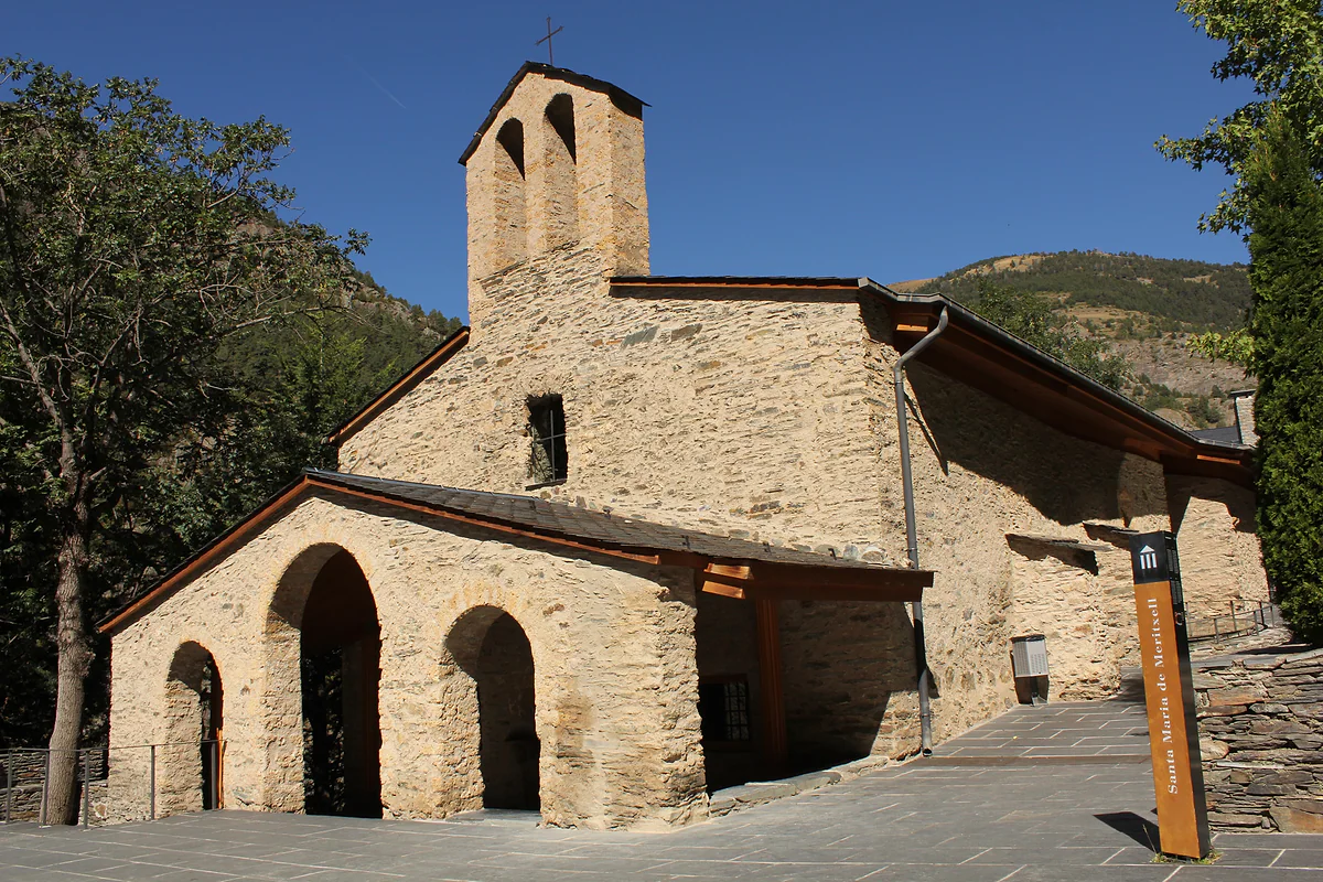 La iglesia antigua de Meritxell, de piedra, con porche de arcos y espadaña, bajo el cielo azul