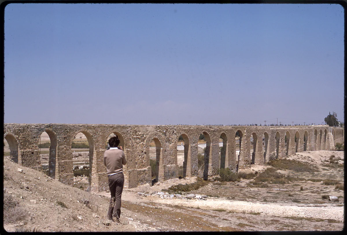 The Kamares aqueduct in perspective, a long row of stone arches, with someone walking along the dirt track beside it