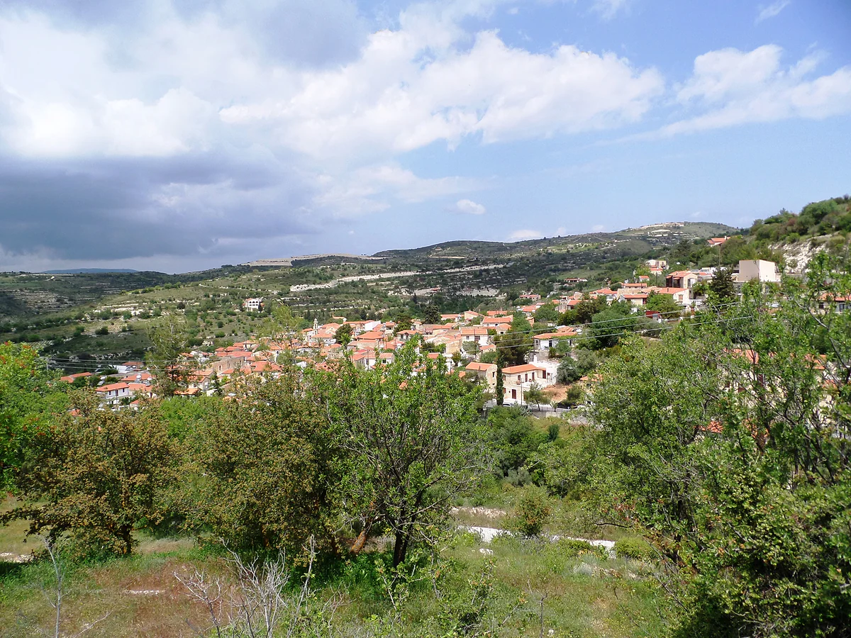 The outskirts of Arsos with trees, scrub and gentle hills, and the village houses peeping out at the far end of the slope