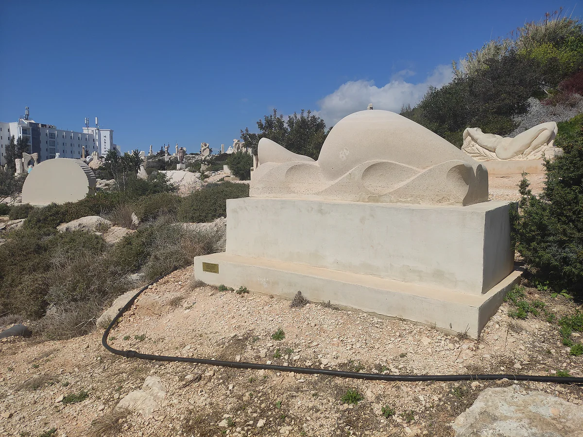 A pale stone sculpture of curving forms on a plinth, on a scrubby slope of the Ayia Napa sculpture park, with other works behind