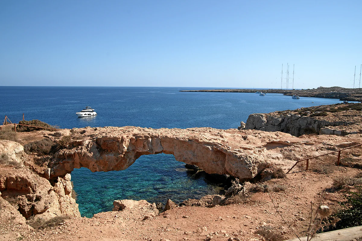The natural arch of Kamara tou Koraka at Cape Greco, spanning a clear pool, with a boat and the open sea behind it, in Cyprus