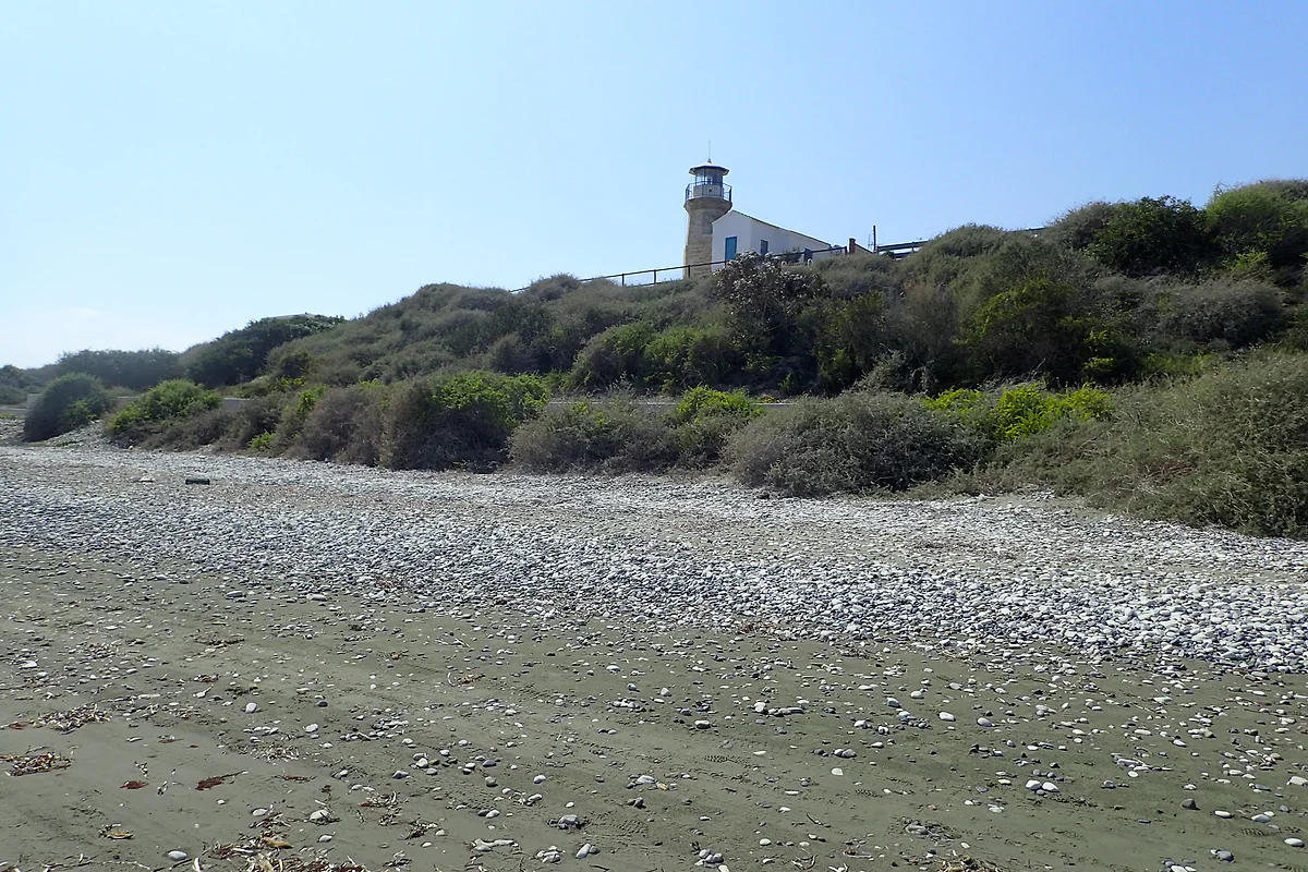 The Cape Kiti lighthouse on a scrubby rise, with a beach of pebbles and grey sand in the foreground under a blue sky