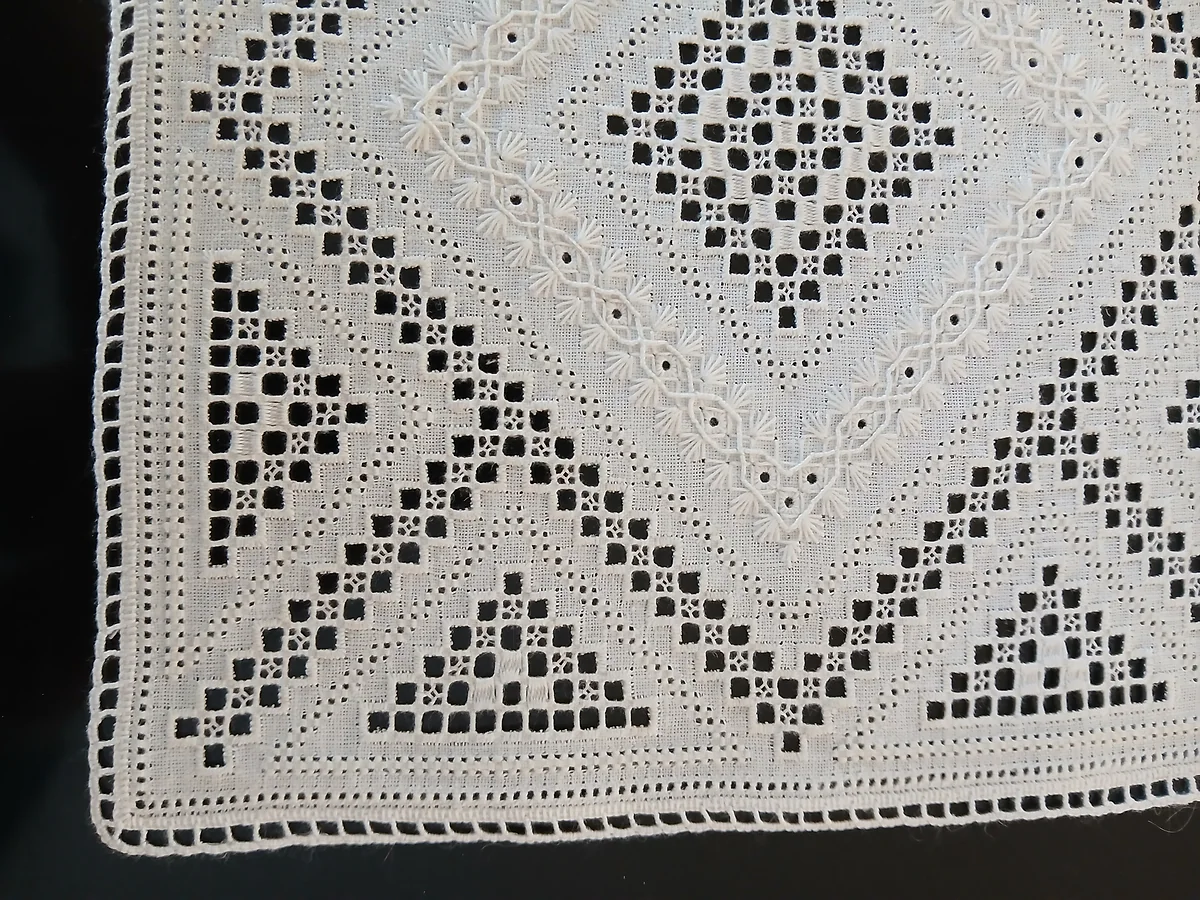 The corner of a handmade piece of Lefkara lace, with white geometric openwork motifs on linen cloth