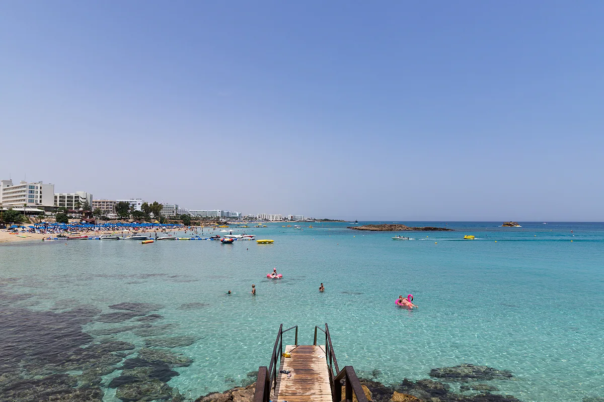 A wooden ladder leads down into the turquoise water of Fig Tree Bay, with swimmers, the islet and the hotels of Protaras across the bay