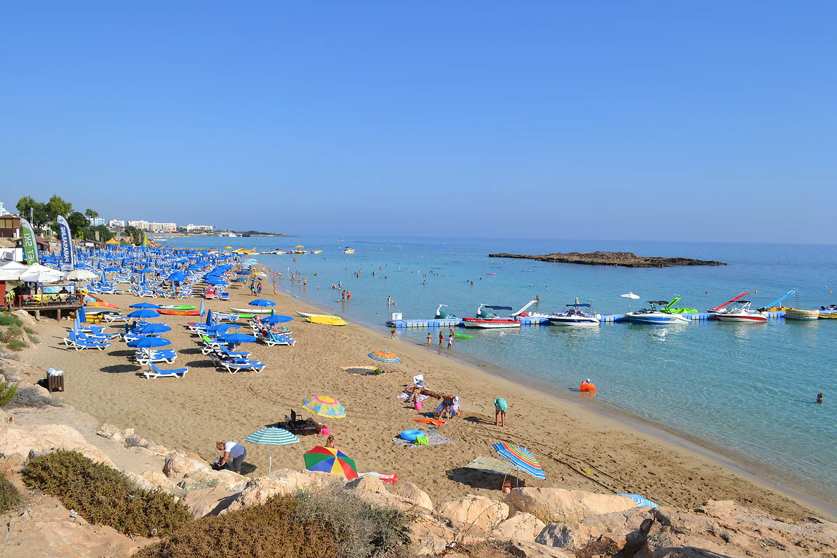 Fig Tree Bay beach in Protaras, with parasols, boats along the shore and the small islet in the middle of the clear water of the bay
