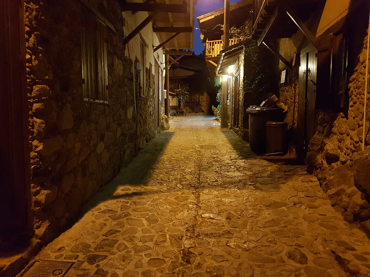 A stone alley in Kakopetria at night, lit by street lamps between old houses with overhanging wooden balconies