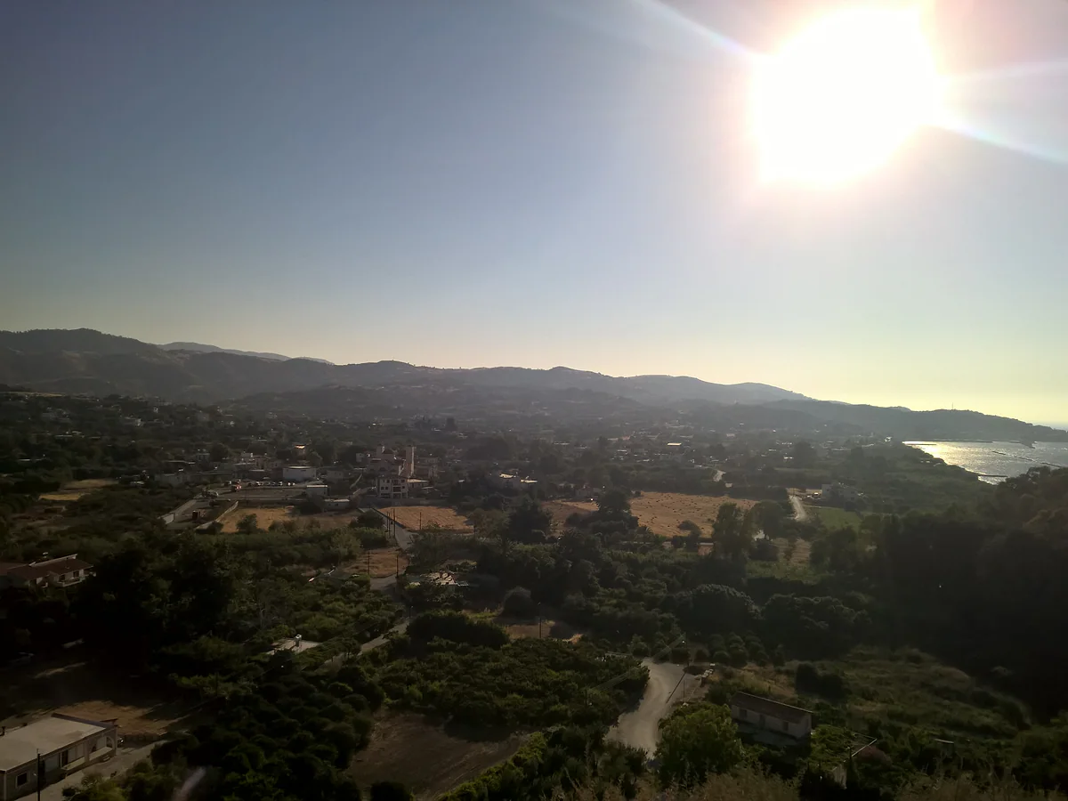 Kato Pyrgos seen from above, the village among orchards and pine woods with the bay behind it against the light, on the Tilliria coast