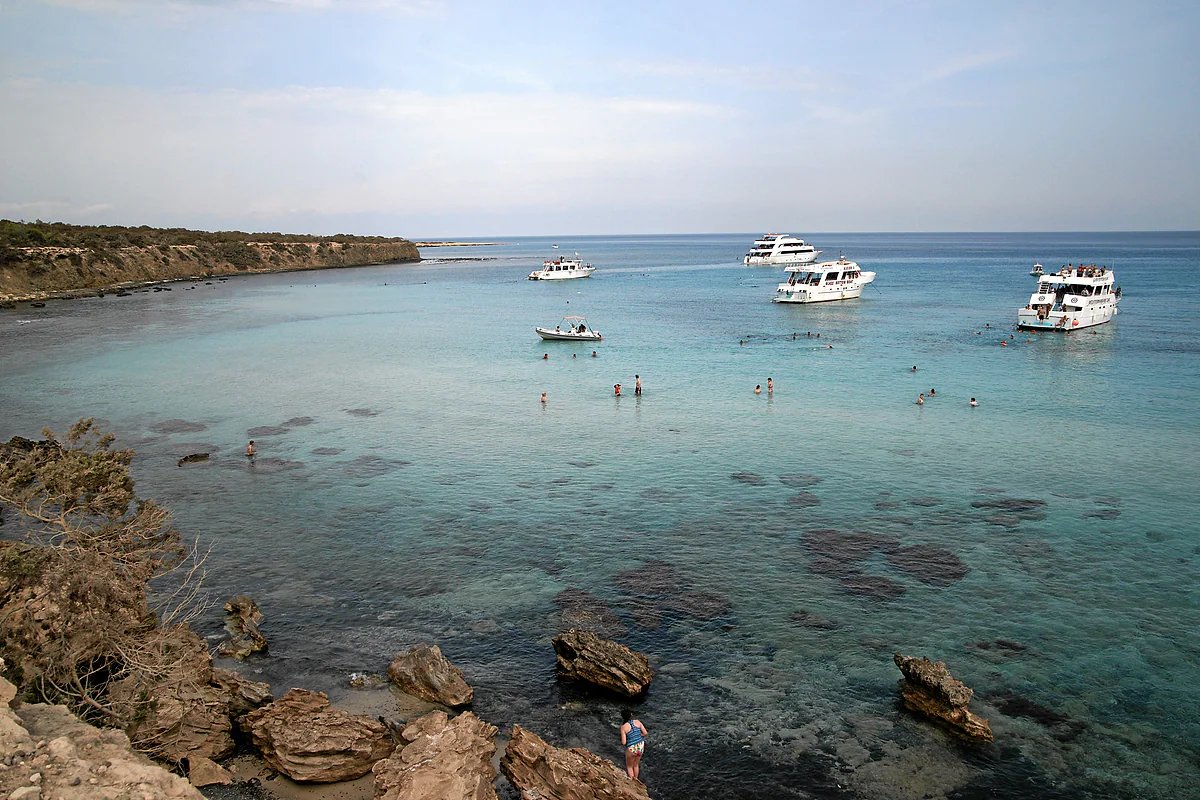 Excursion boats at anchor in the Blue Lagoon off Akamas, with swimmers in the clear water and the pine-clad headland behind