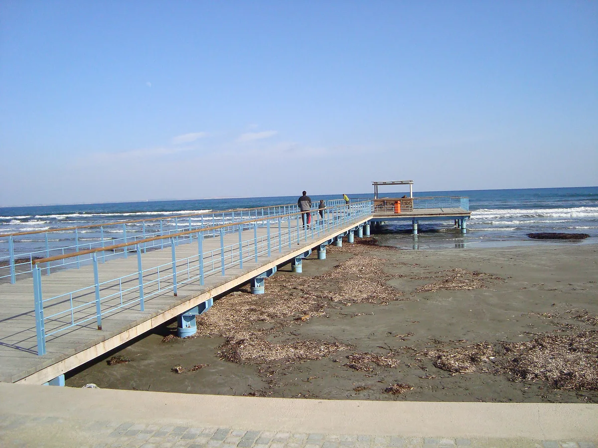 The wooden pier reaching into the sea beside Finikoudes promenade in Larnaca, with seaweed on the sand and gentle waves