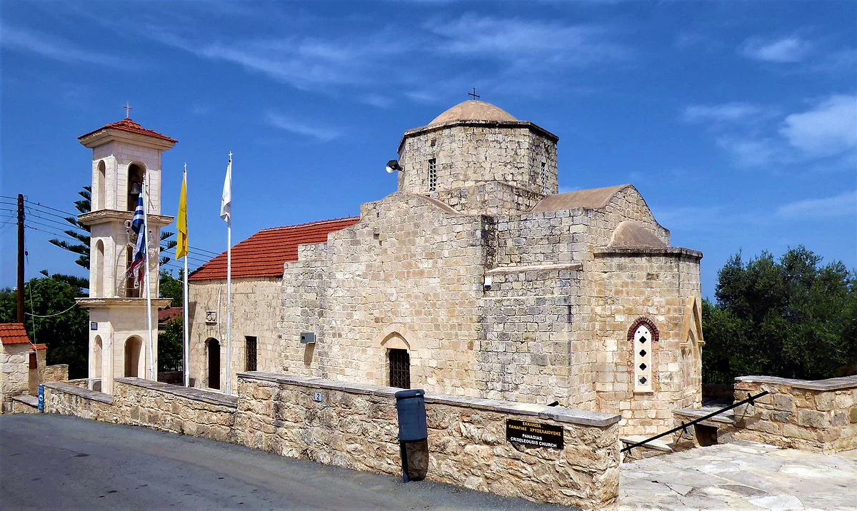 The stone church of Panagia Chryseleousa in Lysos, with its dome and red roofs and a multi-tiered bell tower flying flags