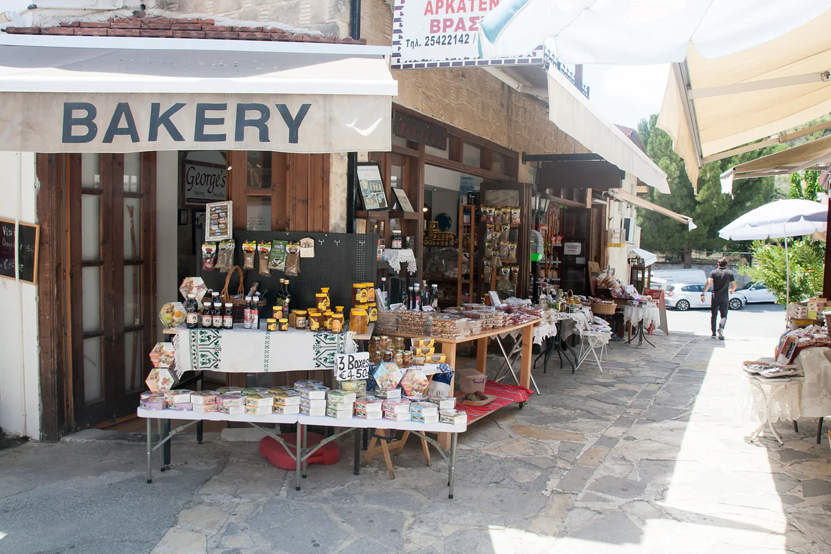 A paved street in Omodos with a bakery and stalls selling sweets, wine and souvenirs beneath canvas awnings