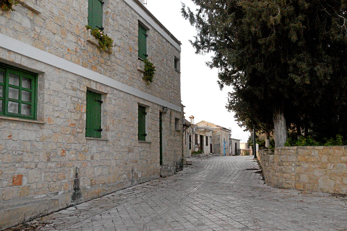 A paved lane in Pano Arodes with two-storey stone houses and green shutters, beside a tall cypress tree in the Paphos hills