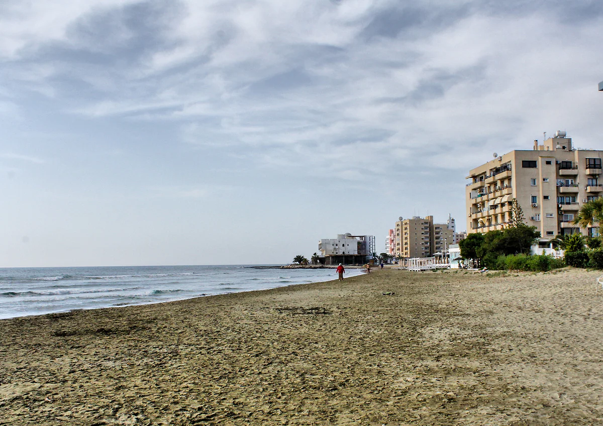 The darker sandy shore of Mackenzie beach in Larnaca, with a calm sea and blocks of flats in the background