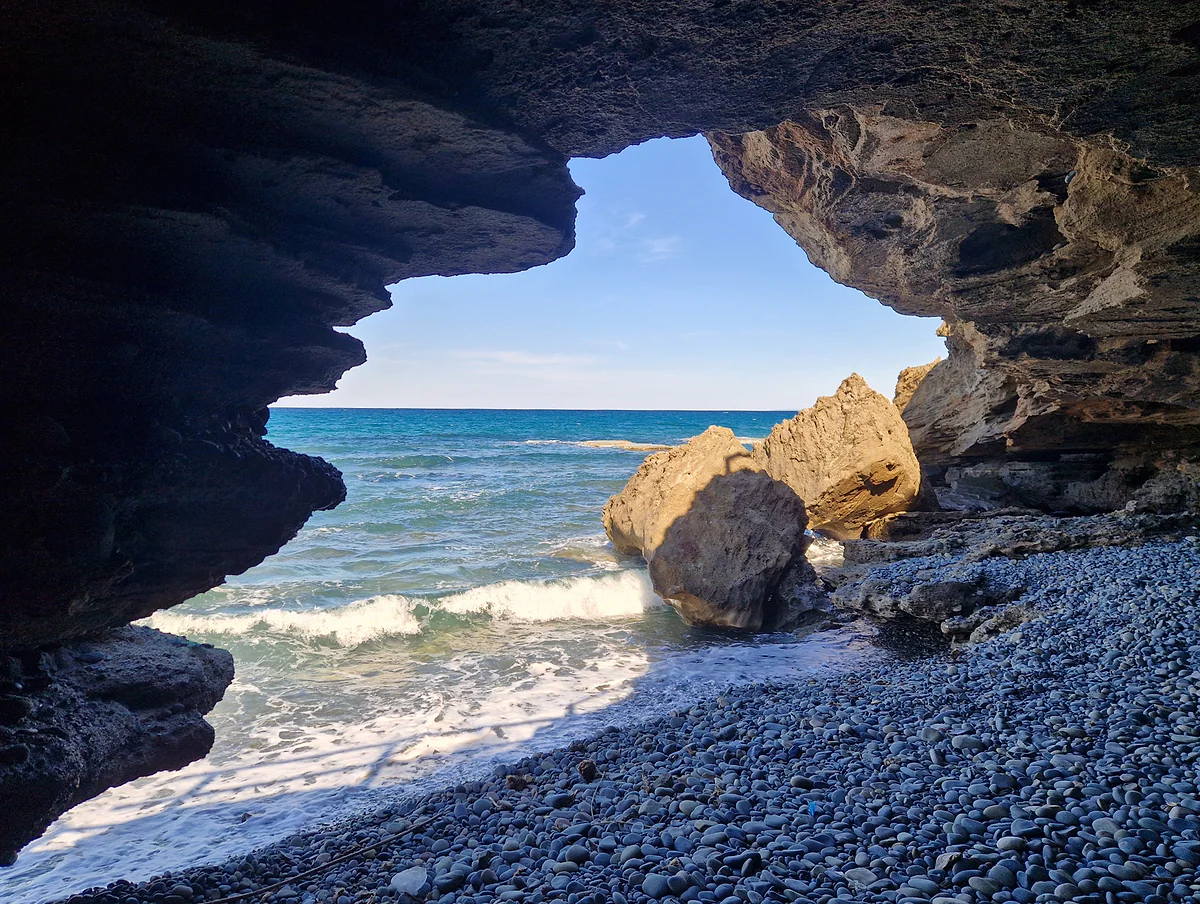 The Dragon's Cave at Pomos, an arch of dark rock open to the sea, with a pebble beach and large boulders at the water's edge