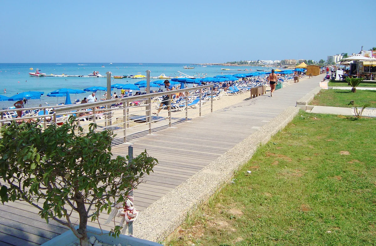 The pedestrian seafront promenade in Protaras, a boardwalk beside a strip of lawn, with the beach and its blue parasols alongside