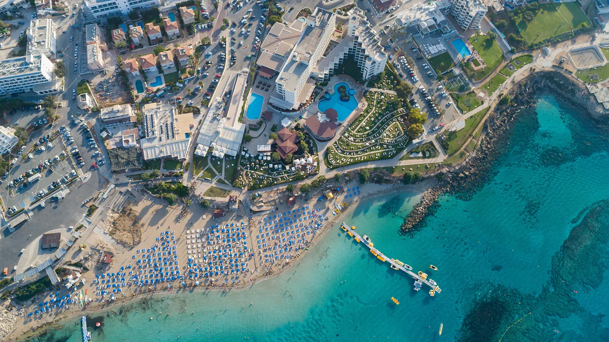 A Protaras beach from the air, with rows of parasols, a floating jetty, hotels with pools and turquoise water