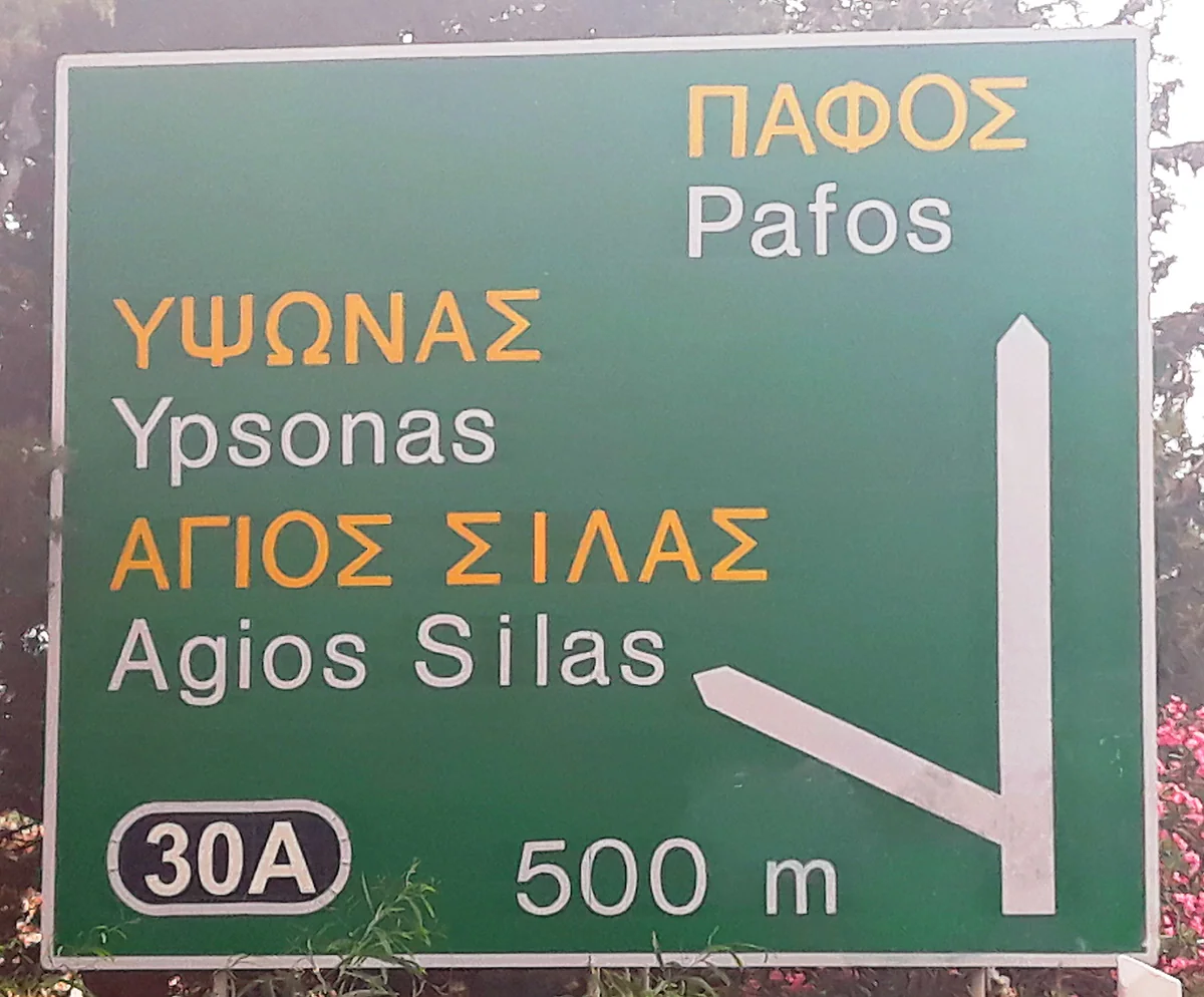 A green motorway sign in Cyprus pointing to Paphos, Ypsonas and Agios Silas, with the names in Greek and Latin script