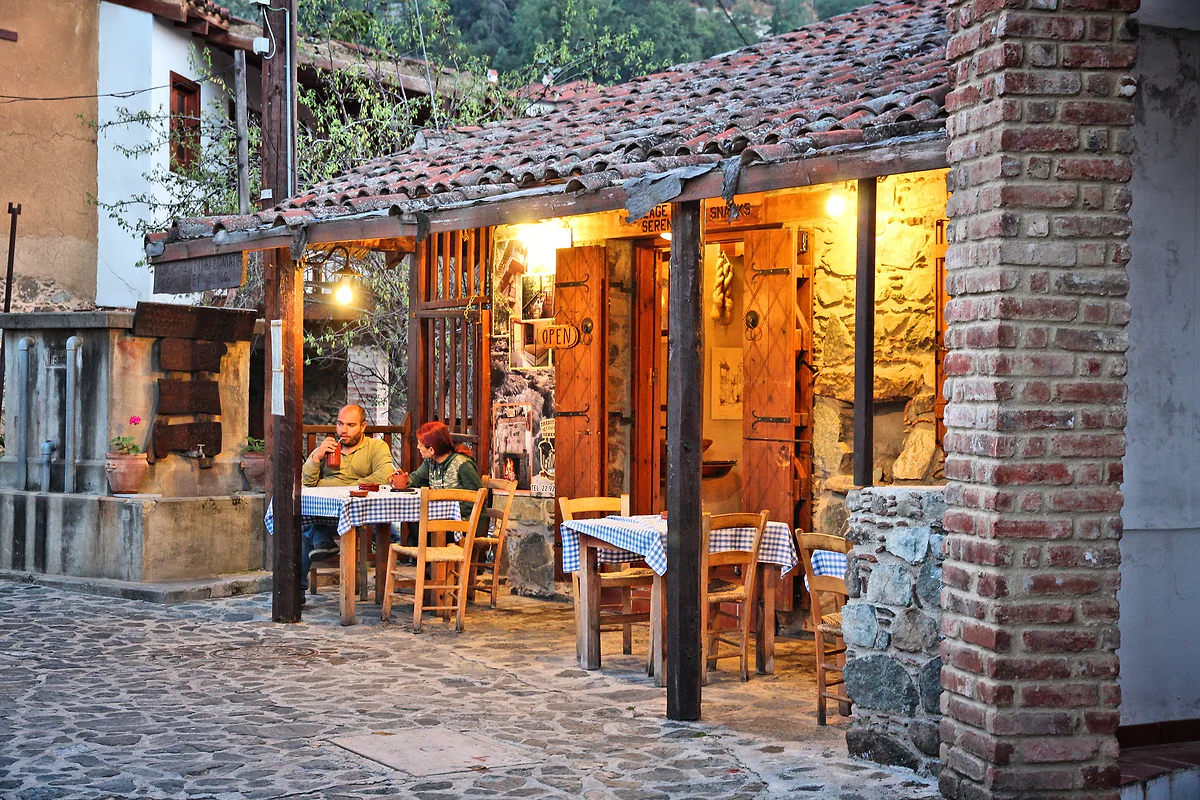 A traditional tavern in Moutoullas with a terrace under a small tiled roof, stone walls and lights on at dusk