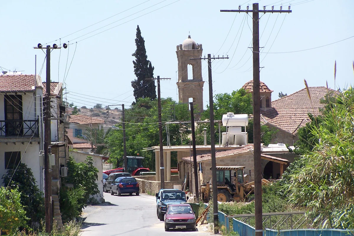 A street in Tochni with parked cars, low houses and the church bell tower rising among the cypress trees