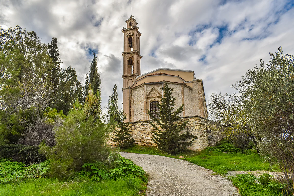 A stone church with a tall bell tower in Vavla, surrounded by cypresses, olive trees and green grass under clouds