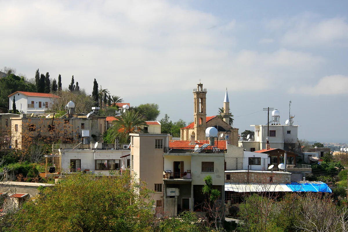 Houses in Lapithos (Lapta) among cypresses and palms, with a church bell tower and a minaret rising above the rooftops