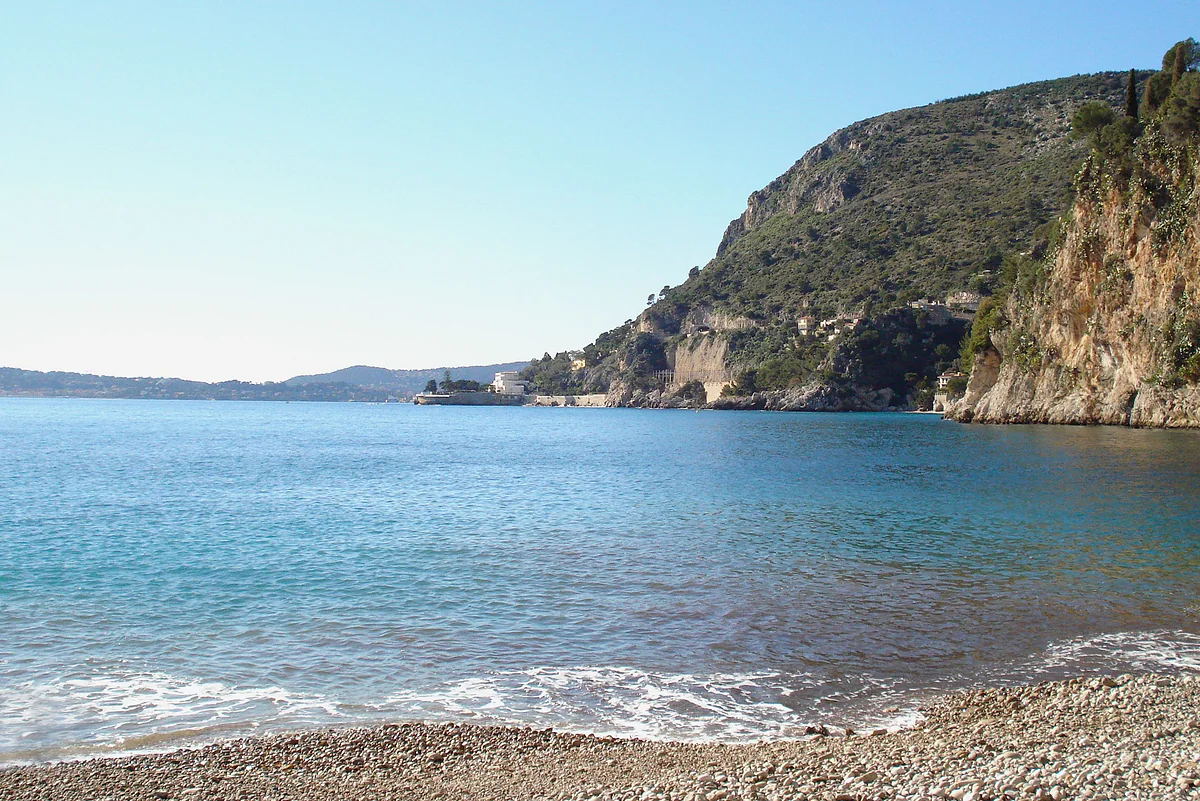 La playa Mala de Cap-d'Ail, de guijarros, con agua turquesa en una cala rodeada de acantilados y el cabo al fondo.