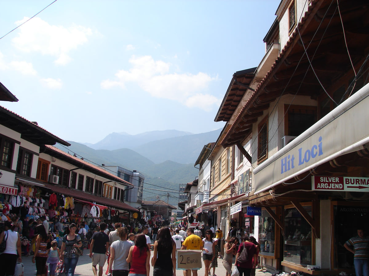 The bazaar street in Peja lined with wooden shopfronts on both sides, people strolling and mountains beyond