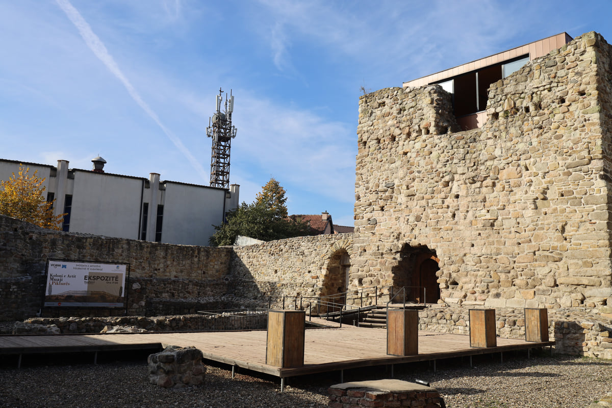 The stone tower of Vushtrri fortress and its low curtain wall, with a wooden stage in the courtyard and town-centre buildings behind