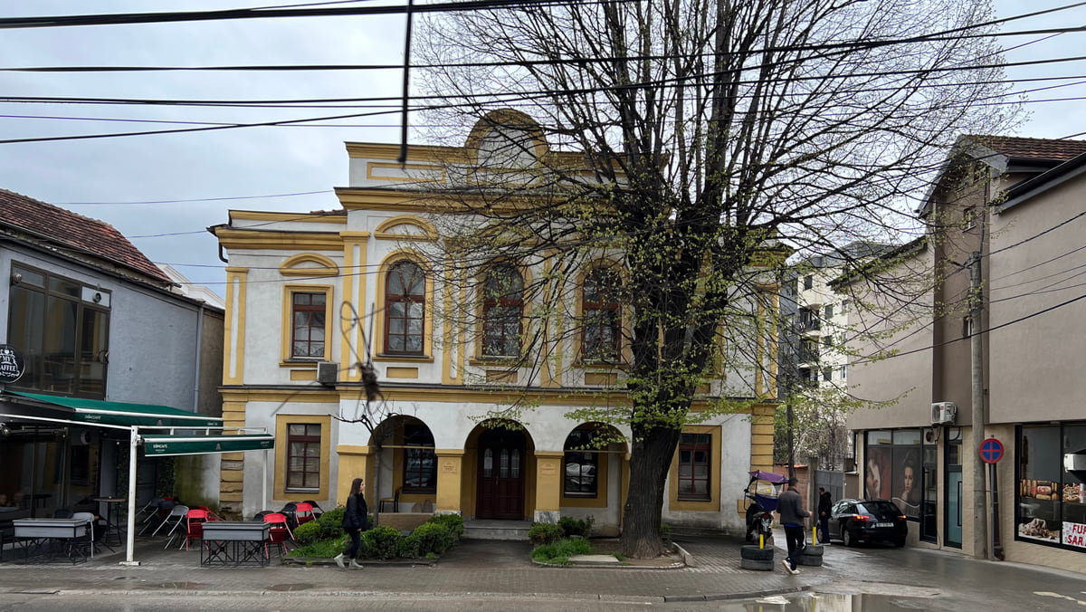 The old Kajmekam building in Gjilan, two storeys in white and yellow with arches, a tree in front and cables over the street