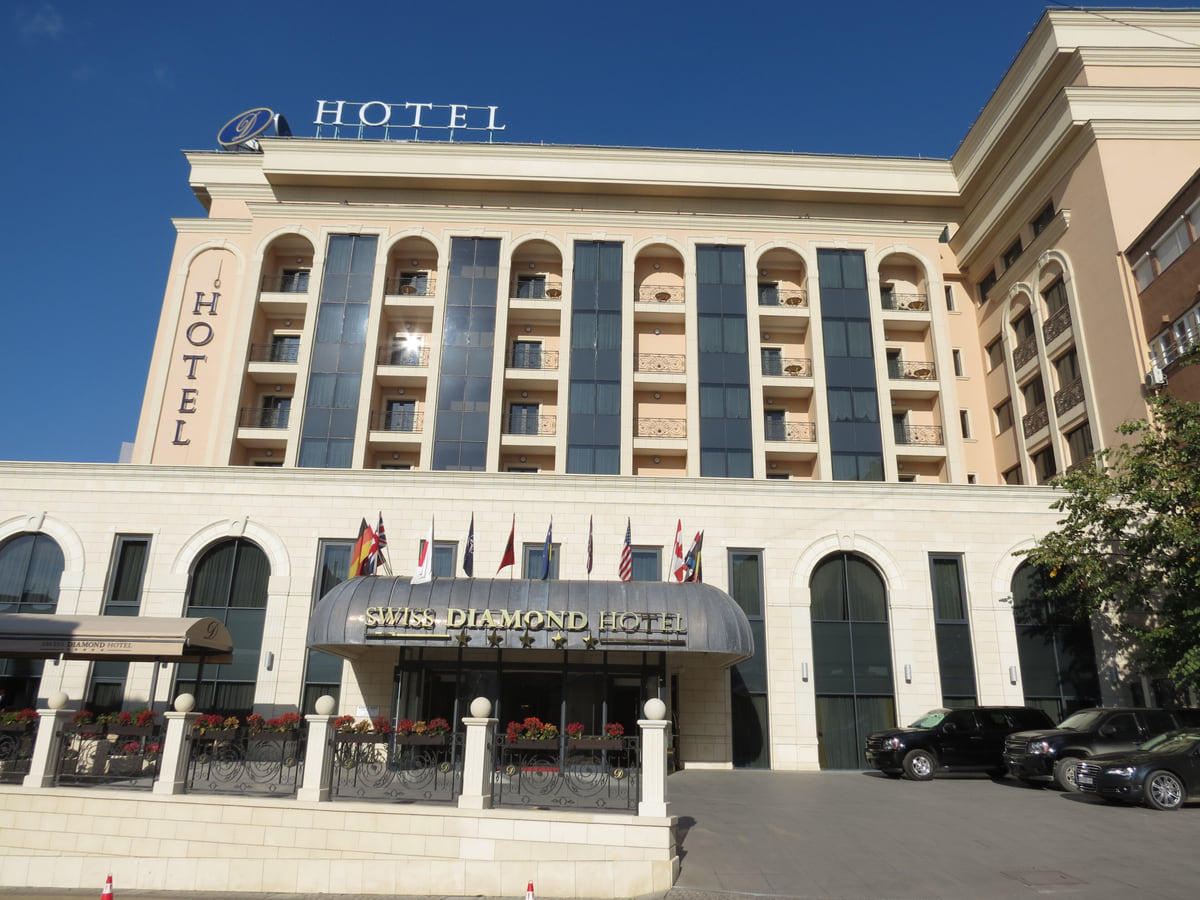 The pale facade of the Swiss Diamond Hotel in Pristina, with its curved canopy, flags and arched balconies under a blue sky