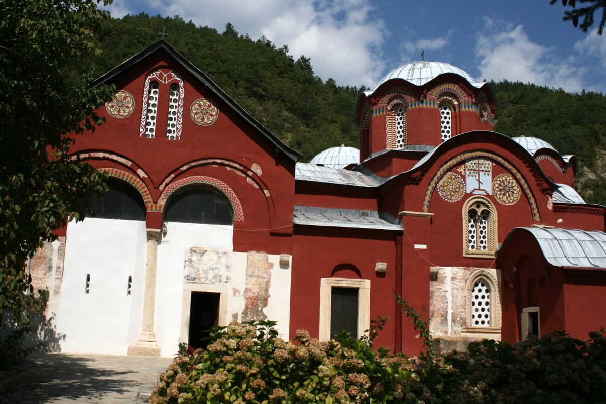 The red facades and domes of the church complex of the Patriarchate of Peć, among trees under a cloudy sky