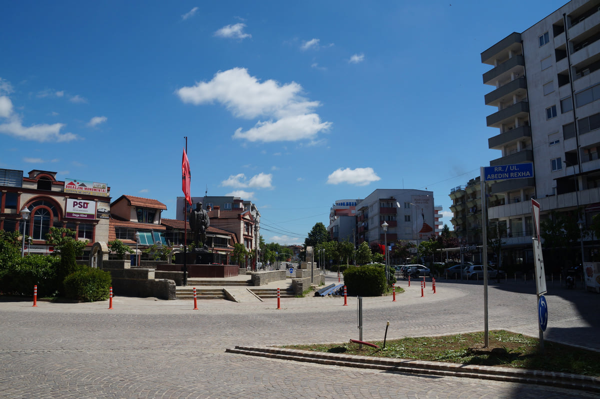 The centre of Klina with a planted roundabout, a flagpole, mid-rise buildings and a blue sky with white clouds