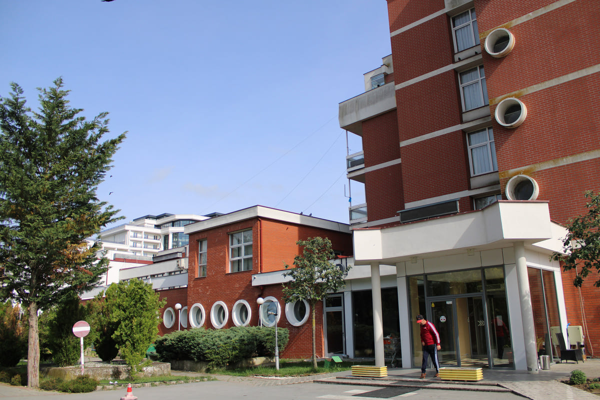 The red-brick Banja building at Kllokot, with round porthole windows, a glazed entrance and conifers in front