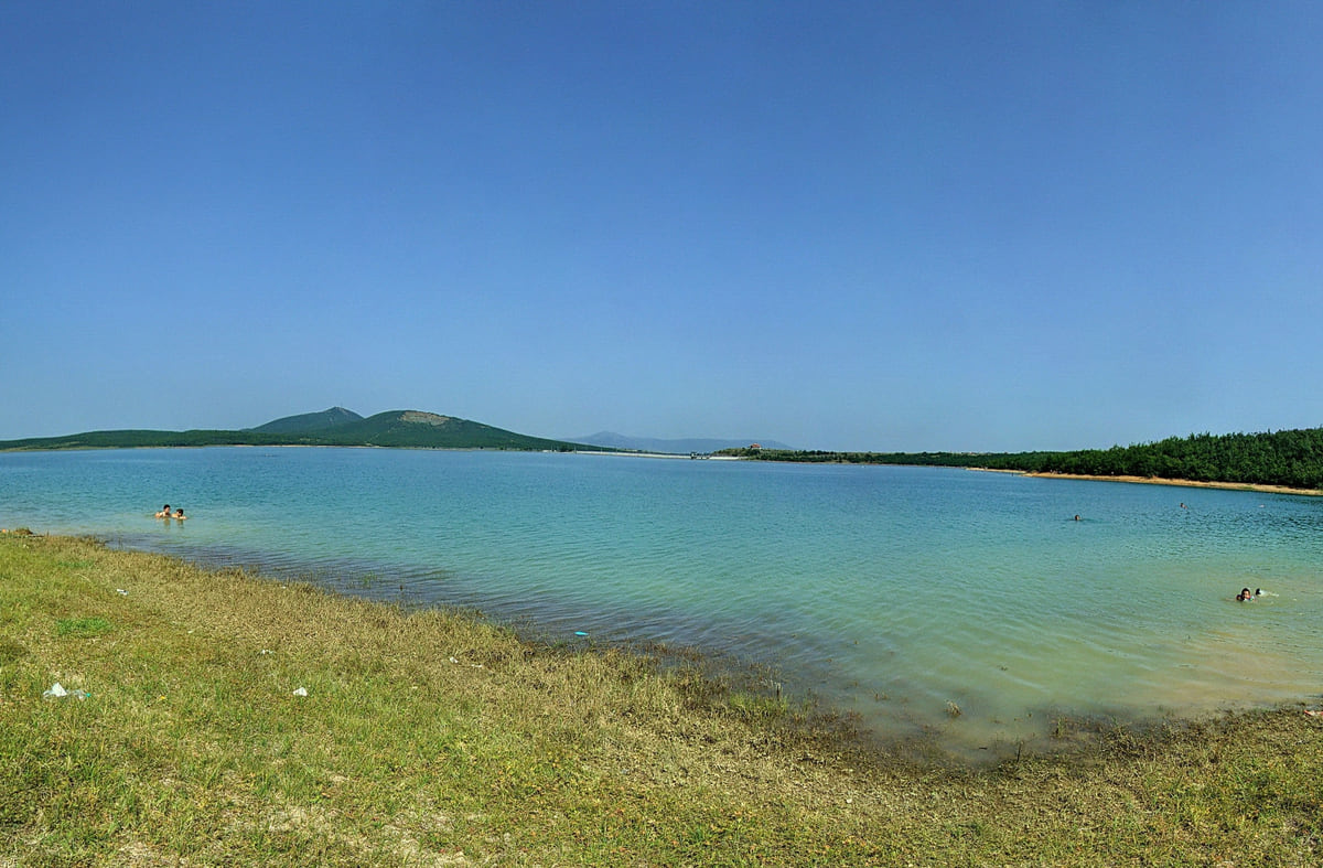 The shore of Lake Radoniq with grass in the foreground, pale blue water and low hills beyond under a clear sky