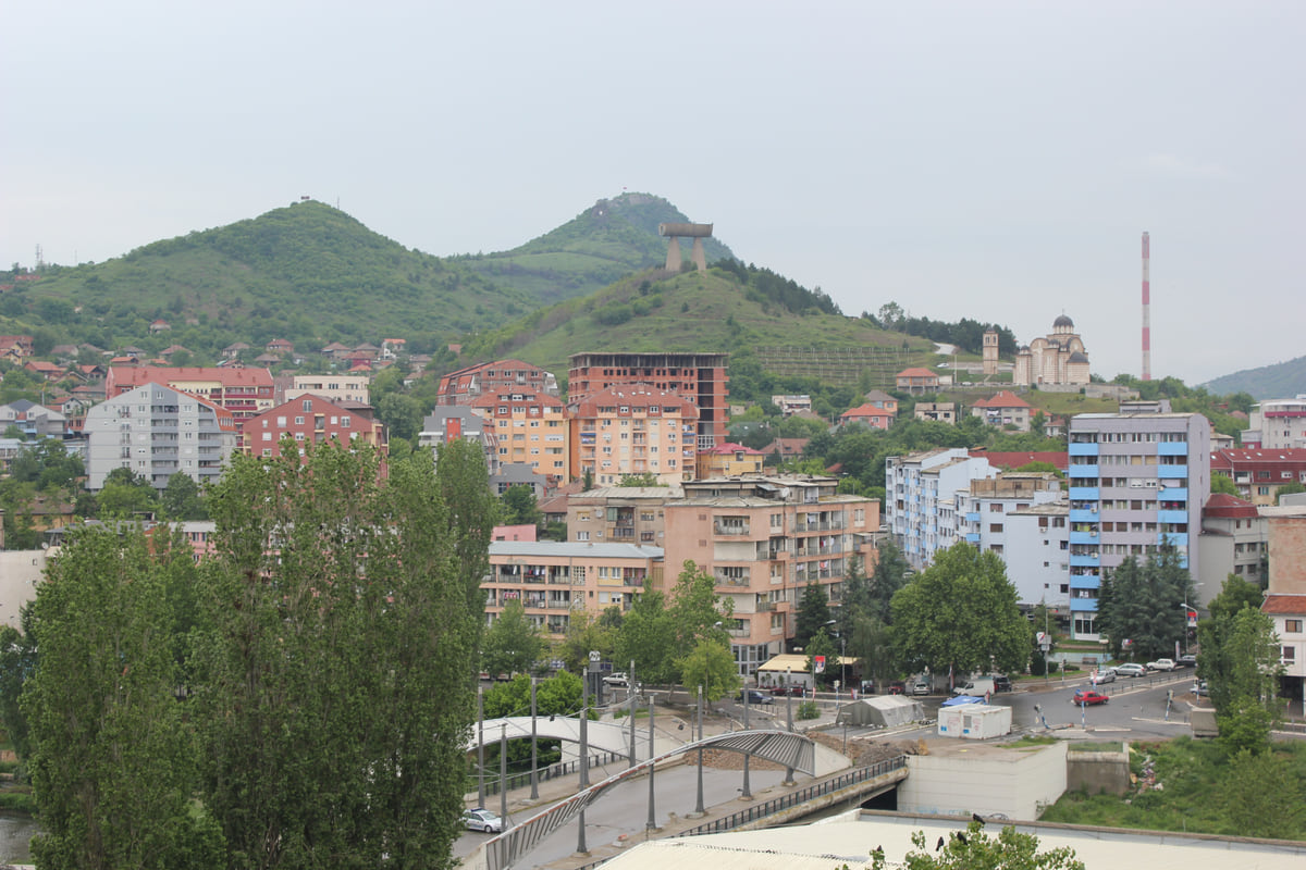 The northern part of Mitrovica seen from above, with the footbridge over the Ibar, blocks of flats and green hills behind