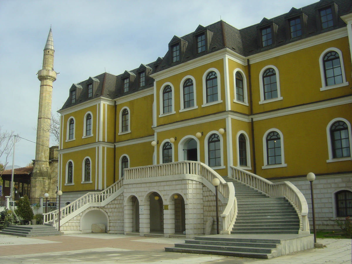 The yellow building of the Kosovo Museum in Pristina, with its white double staircase and a minaret rising behind it