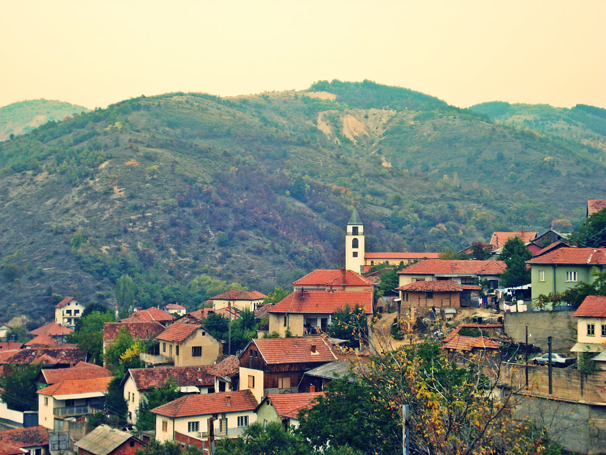 The village of Janjevo (Janjevë) on a hillside, with red-roofed houses, the church tower and green hills behind