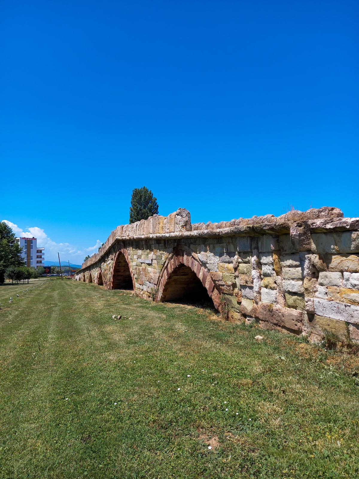 The multi-arched stone bridge of Vushtrri (Vučitrn) standing on grass under a deep blue sky