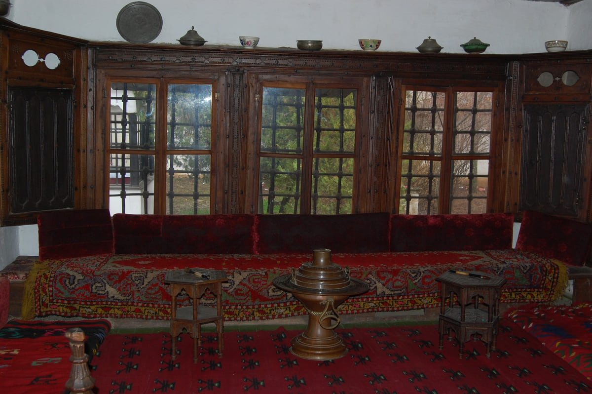 A room in the Ethnological Museum in Pristina with red rugs, low cushions along the walls and latticed windows