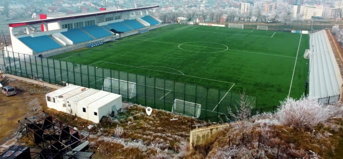 Shtime's football ground seen from above, with green pitch, blue stands and the town behind dusted with frost
