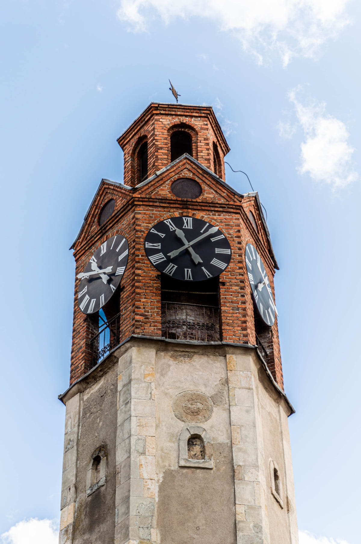 The brick clock tower of Pristina with white clock faces and an iron lantern on top, seen from below against a clear sky