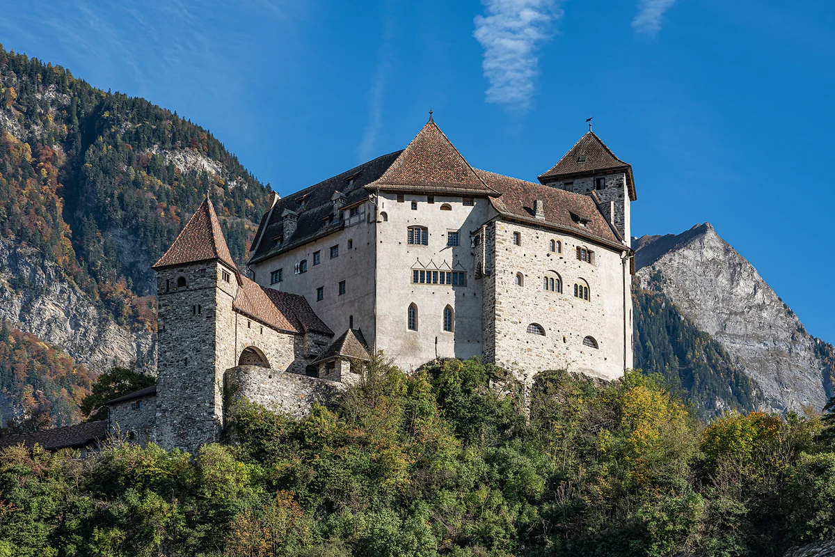 El castillo de Gutenberg en Balzers, con sus torres y muros blancos sobre la colina boscosa y una montaña rocosa detrás.