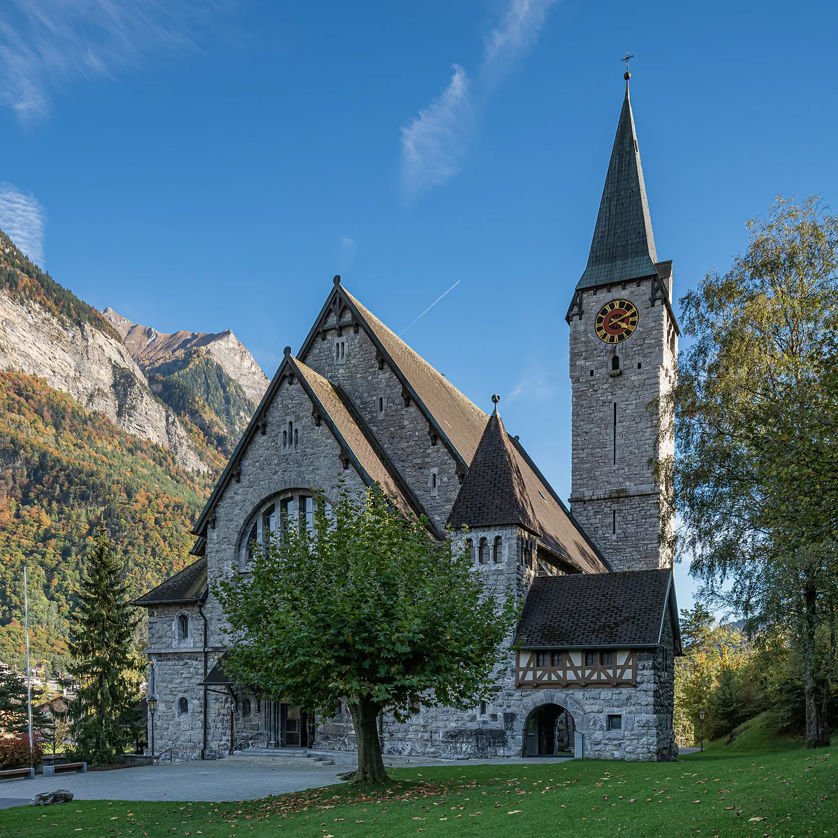 La iglesia parroquial de San Nicolás de Balzers, de piedra gris y campanario puntiagudo, con los montes al fondo.