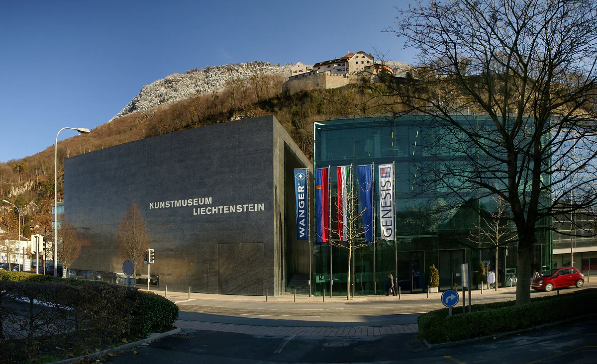 El cubo negro del Kunstmuseum Liechtenstein junto a la calle, con las banderas en la entrada y el castillo en lo alto de la roca.