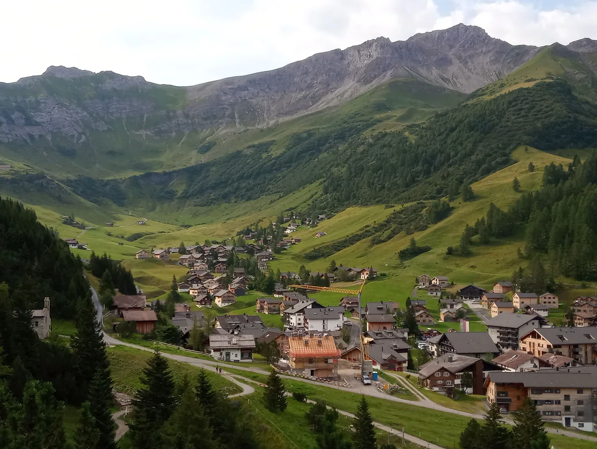 Malbun en verano, con el pueblo de chalés en el fondo del valle, los prados verdes y las cumbres rocosas encima.