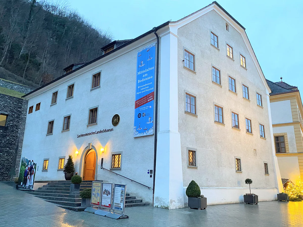 La fachada blanca del Museo Nacional de Liechtenstein al atardecer, con la puerta iluminada y un cartel de exposición.
