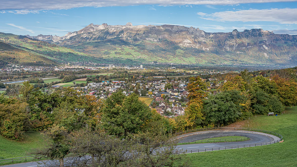 El valle del Rin visto desde el castillo de Vaduz, con la ciudad entre árboles otoñales y los montes suizos en el horizonte.