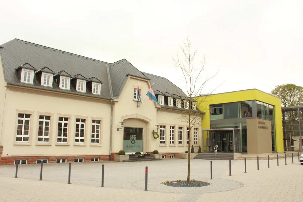 Rambrouch town hall, a pale building with slate dormers beside a modern yellow extension and a small square