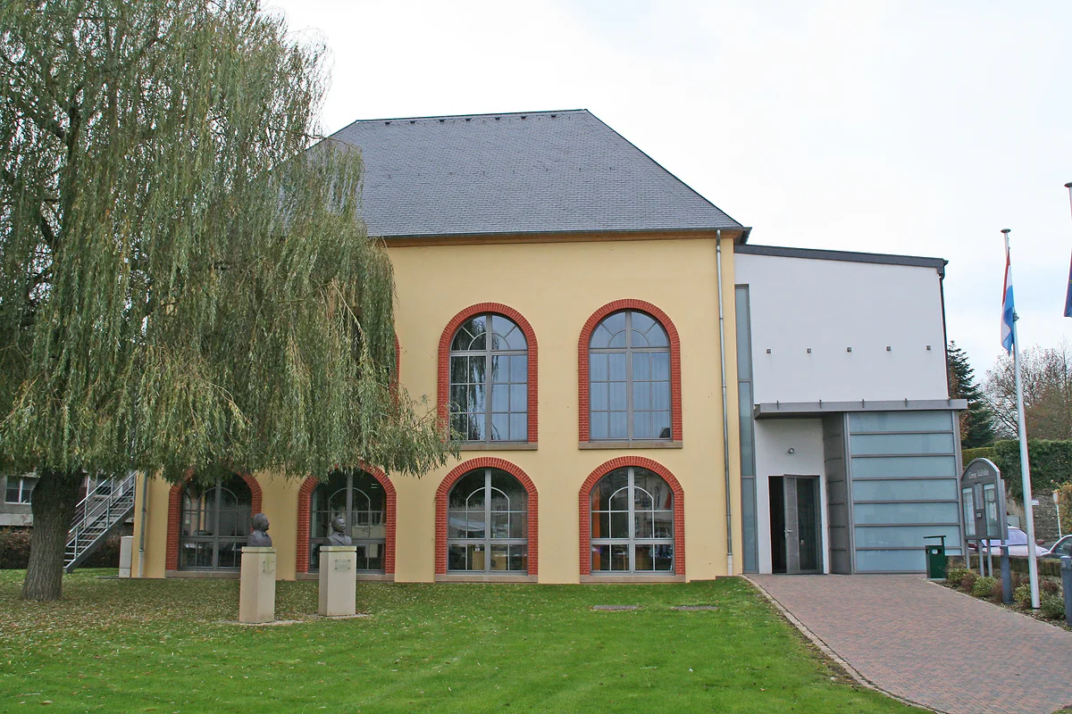 Waldbillig town hall, with large arched windows framed in red, a weeping willow and a lawn in front
